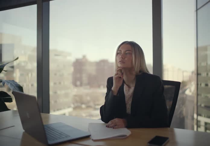 Woman sitting at an office table with a laptop computer as she fills out paperwork and stops to think