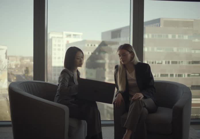 Two women sitting side by side in a highrise office building at midday discussing as one shows the other a laptop screen