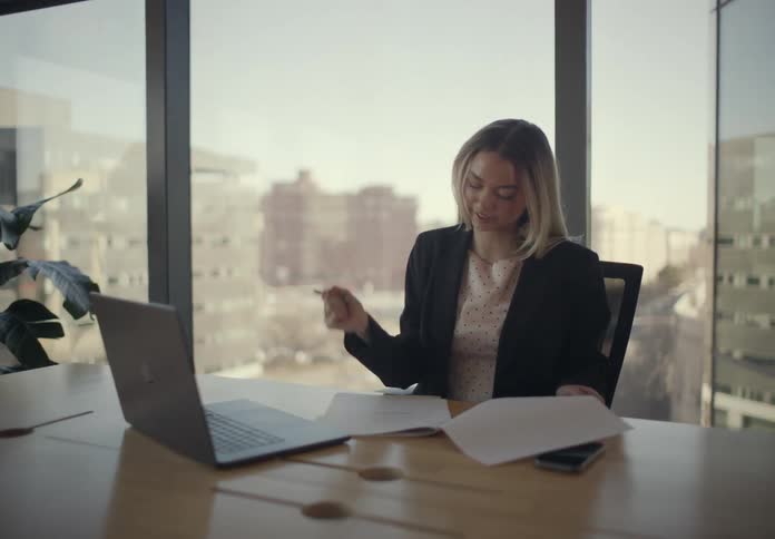 A blonde woman in a blazer sits at a modern desk by a large window reviewing documents and writing notes with a pen while a laptop and phone remain stationary nearby