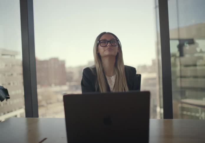 A professional woman sits at a desk in a modern office typing on her laptop while looking thoughtfully out the window at the city she eventually stops working closes her laptop and leans back in her chair with a calm accomplished expression