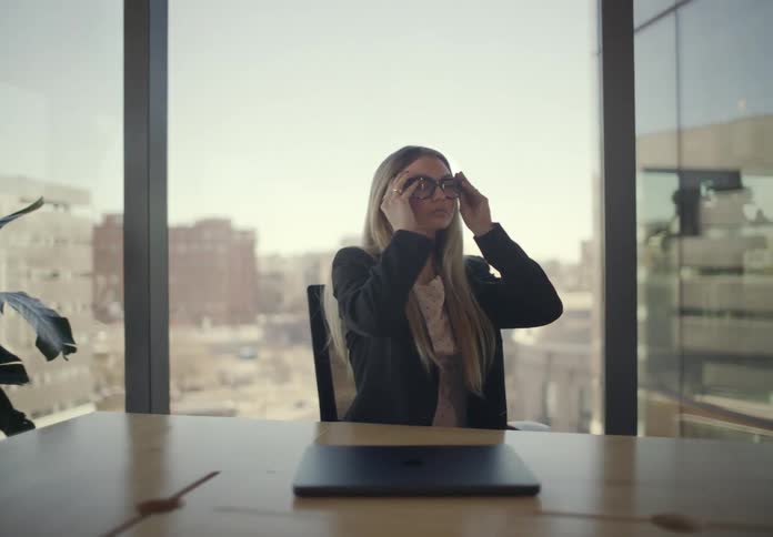A young woman wearing glasses sitting at a desk at midday closing the lid of the laptop in front of her