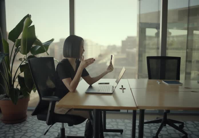 Woman talking on a smartphone video call while working at a meeting table with a laptop and chairs around