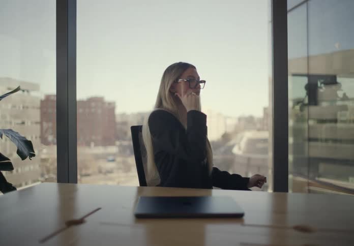 A woman in a black blazer closes her laptop at a conference table and puts on her glasses while looking out a large office window the scene overlooks a city view under bright daylight