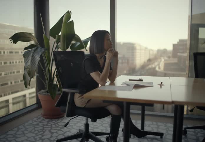Young businesswoman in a modern sunlit office sitting at a desk with papers looking thoughtfully before leaning forward to write with a city view in the background