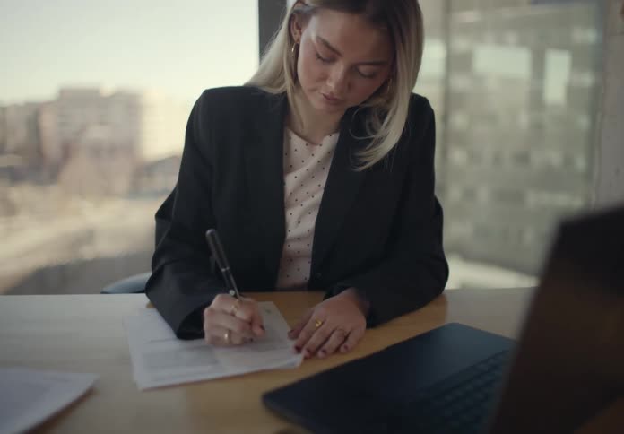 A young woman with long blonde hair wearing a blazer suit sitting behind a desk in a highrise office writing on a document with a laptop in front of her and a large glass window behind her