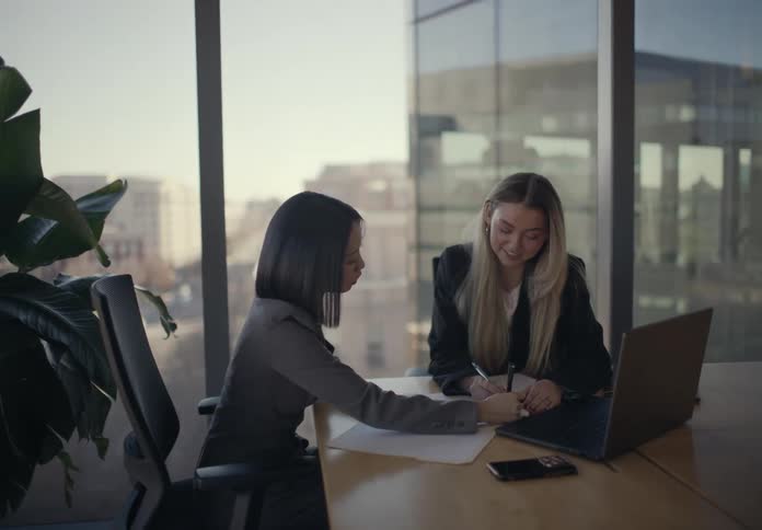 Two women reviewing documents at a meeting table with an open laptop and glass windows in the background
