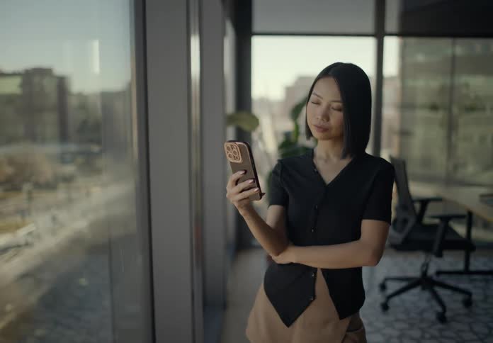 Young adult female using a smartphone standing up in a room with big windows showing a metropolitan area indoors during day time