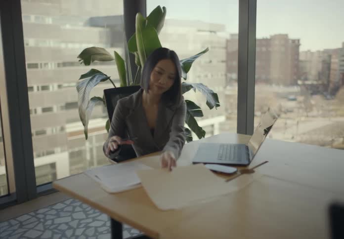A woman walks across a modern highrise office to hand a folder to a colleague seated at a desk then the seated woman then reviews the documents and begins writing next to a laptop displaying a colorful pie chart