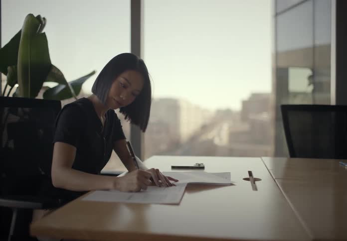 A woman writes on documents while seated at a conference table in a bright modern office