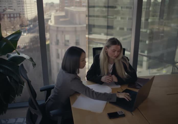 Two women collaborating at a meeting table with documents and a laptop in front of glass windows with an urban view