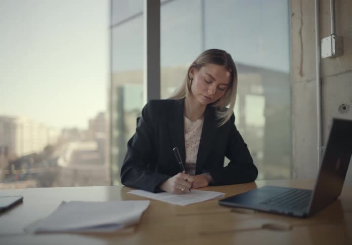 A professional woman sits at a desk reviewing paperwork beside a laptop in a modern office