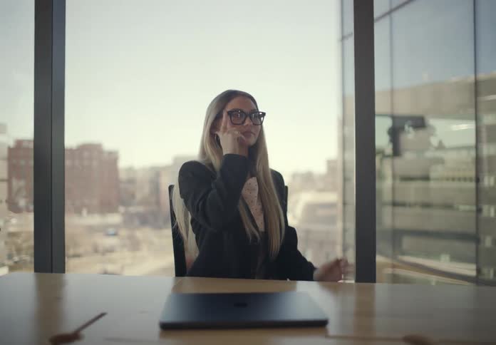 A young woman sits in a modern office and closes her laptop and puts on eyeglasses at a desk