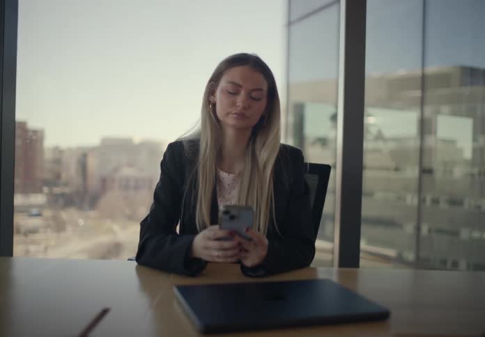 A woman in a professional blazer sits in a bright office thoughtfully scrolling through her smartphone she pauses her browsing to look out the large window at the city view appearing calm and focused