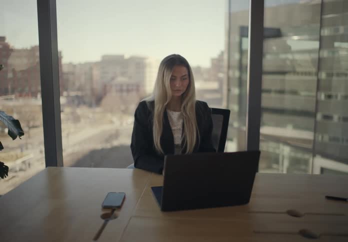 Woman typing on a laptop at a meeting table with a smartphone beside her and an urban view through glass windows