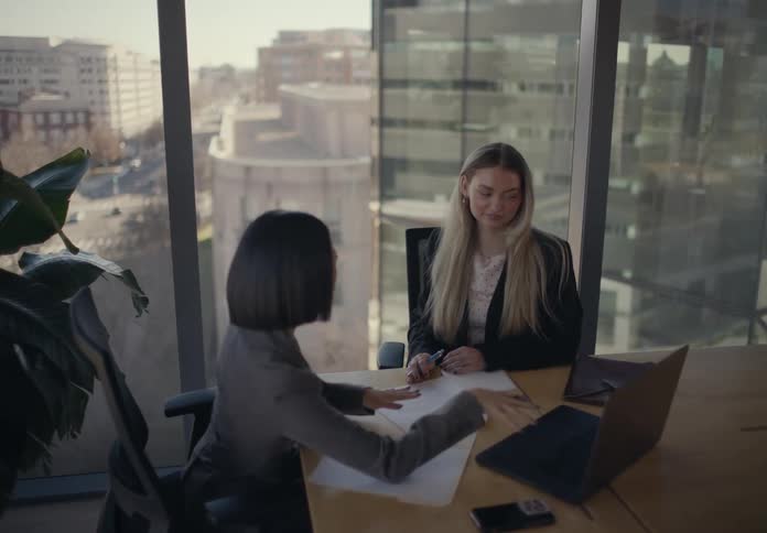 Two professional women sit at a large wooden desk in a modern office collaborating on a project with a laptop and various documents they engage in a friendly discussion while looking at the screen surrounded by expansive windows that offer a clear view