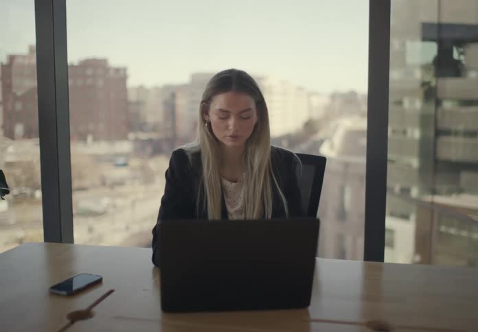 A young female office worker with long blonde hair typing on a laptop inside an office at midday with her mobile device beside her