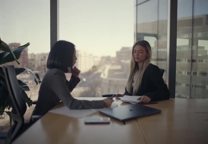 Two professional businesswomen sitting at a desk in a sunlit office engaging in a friendly and animated conversation while reviewing paperwork