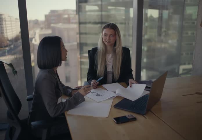 Two professional women sit at a table in a modern office with large windows overlooking a city attentively reviewing and annotating printed documents together while an open laptop a smartphone