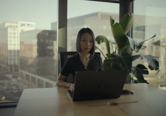 A businesswoman walks into a modern office with large windows sits down at a conference table and begins working on her laptop