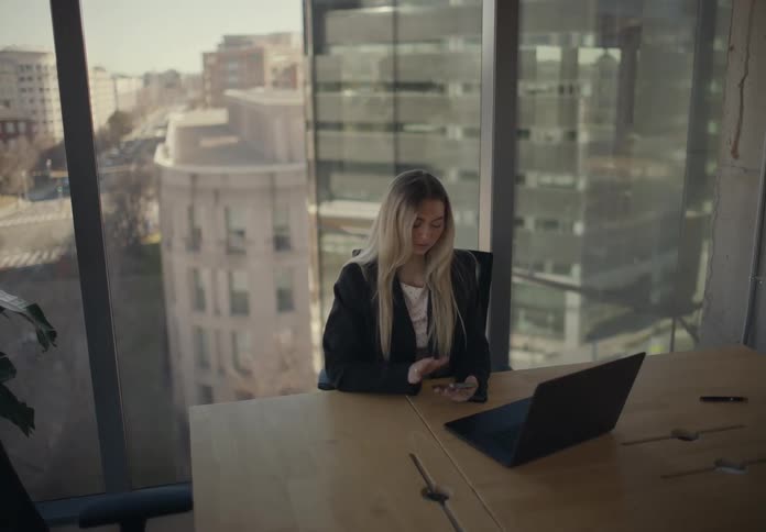 The woman is using her cell phone and laptop at the desk