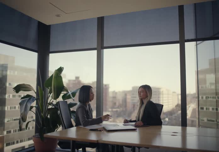 Two women sitting in an office talking over paperwork with a phone and laptop computer on the table