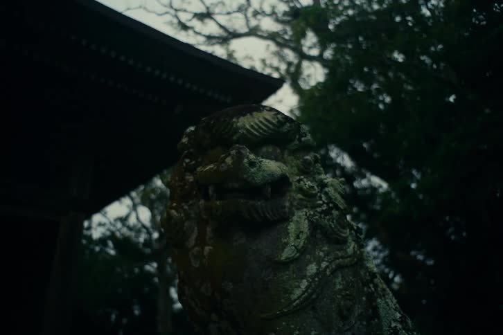 An ancient stone lion statue stands weathered and covered in moss positioned in front of a traditional darkroofed building the scene is set in a dimly lit overcast environment with thick green foliage visible in the background