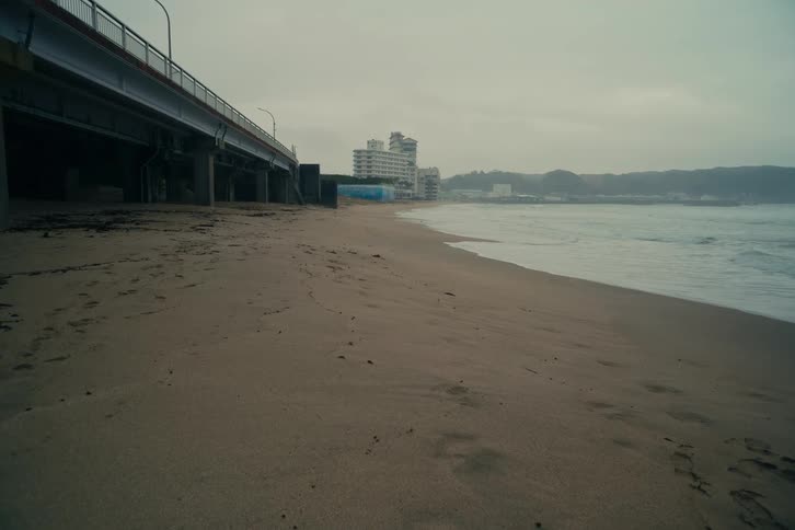 A deserted beach on a gloomy overcast day features a large bridge to the left and a city skyline in the distance