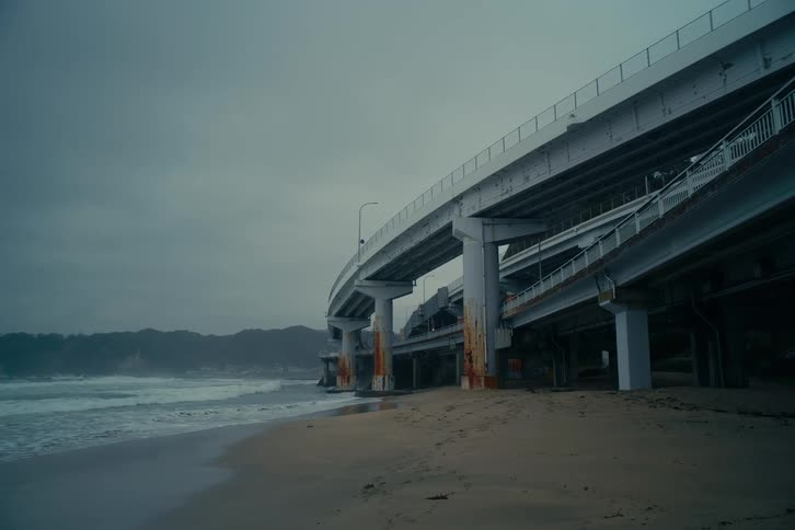 A massive concrete highway overpass looms over a desolate beach coastline beneath a overcast sky
