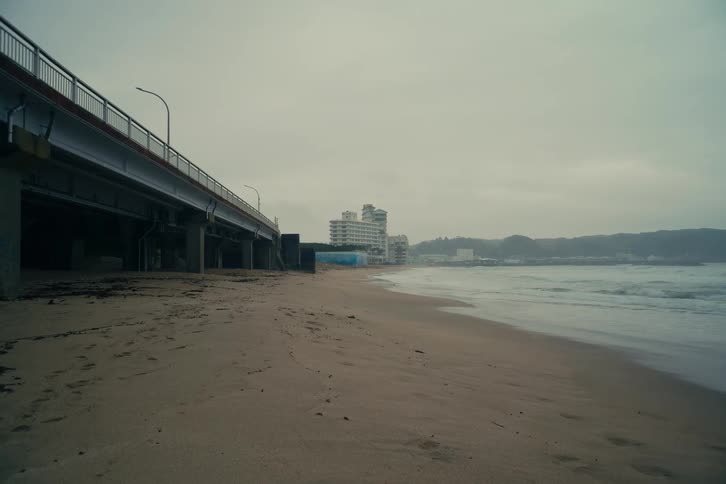 An overhead bridge above a beach on a cloudy day with waves hitting the shoreline
