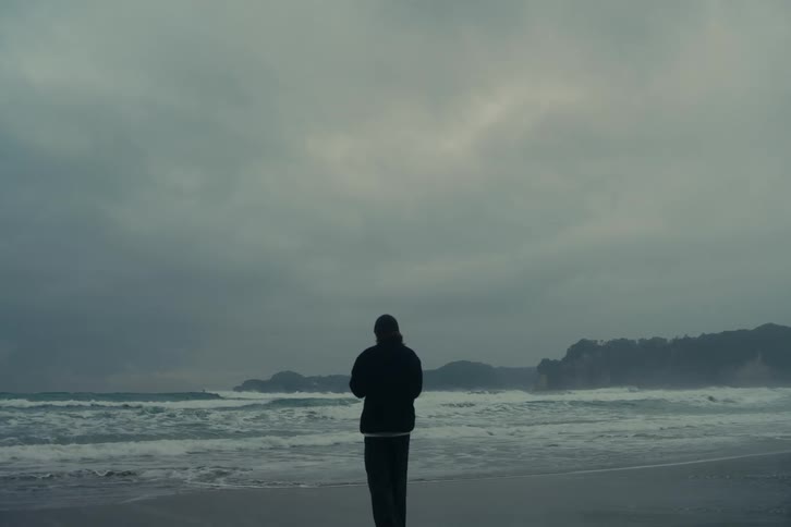 A lone person walks toward the ocean along a quiet windswept beach under a dark overcast sky