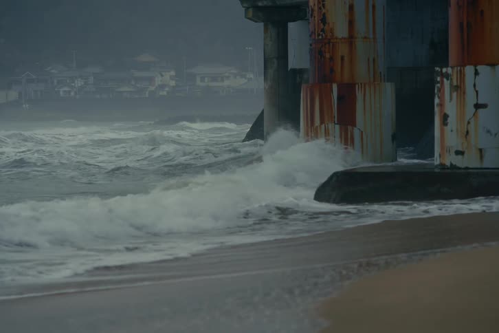 Heavy churning ocean waves slam into the base of massive ruststained bridge pillars along a sandy shore in the background a row of houses and dark misty hills are partially obscured by the salt spray and the dim overcast light of a coastal day