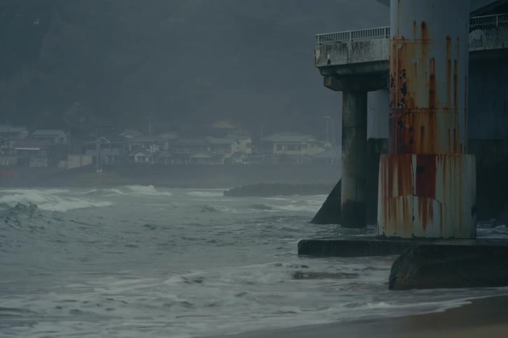 Heavy churning ocean waves slam into the base of massive ruststained bridge pillars along a sandy shore in the background a row of houses and dark misty hills are partially obscured by the salt spray and the dim overcast light of a coastal day