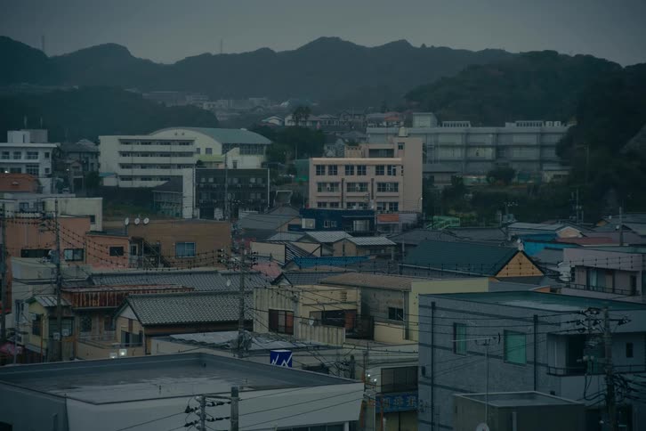 A densely populated residential area in japan during the blue hour characterized by a mix of lowrise houses and larger apartment blocksin the background dark forested hills sit beneath a heavy overcast sky creating a somber and quiet atmosphere a