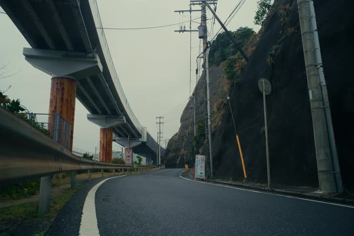 A silver minivan drives along a winding mountain road in japan passing under a massive concrete highway overpass while flanked by a steep rocky cliff and utility poles under an overcast sky