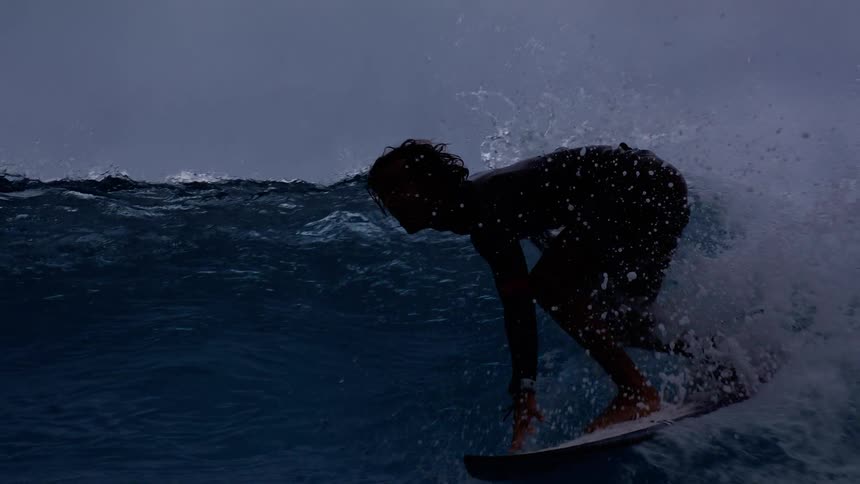 A silhouetted surfer wearing a dark wetsuit rides across a breaking wave under an overcast sky generating white spray before the camera moves into the churning water and foam