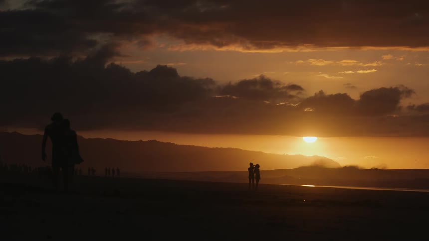 Silhouettes of people walking on the beach at sunset with mountains in the background and clouds in the sky