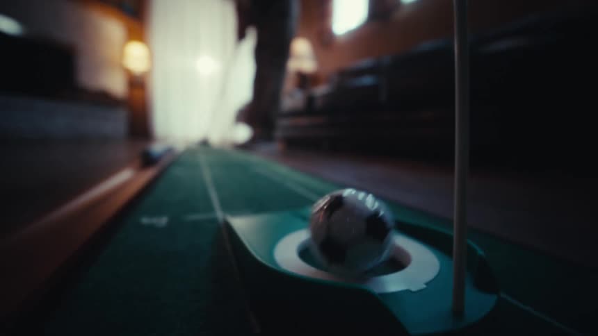 A soccer patterned golf ball rolls across an indoor putting green and into the hole