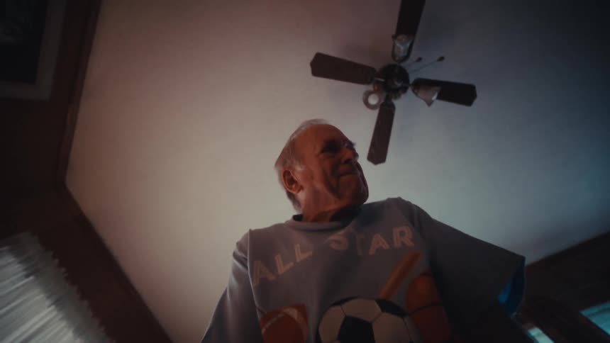 A senior man wearing a soccer tshirt practices his golf putt on a small indoor green inside a living room smiling as he looks down at the ball before the camera angles up toward his face and the ceiling fan above him