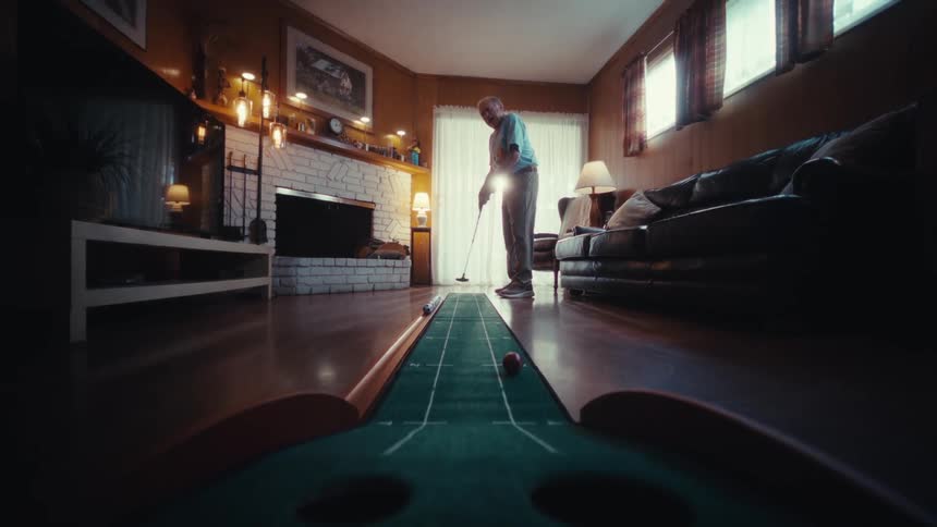 An elderly man in a blue shirt practices his golf putting on an indoor green inside a woodpaneled living room