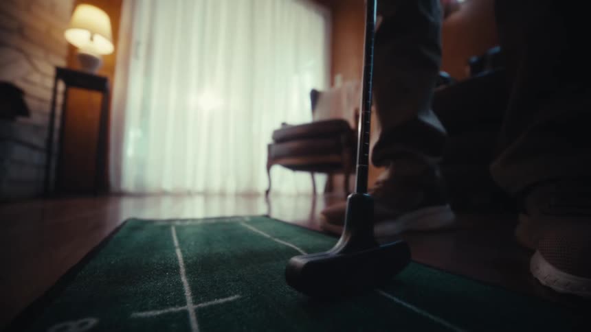 A person uses a black putter to strike a red and white patterned golf ball across a green indoor putting mat in a dimly lit living room