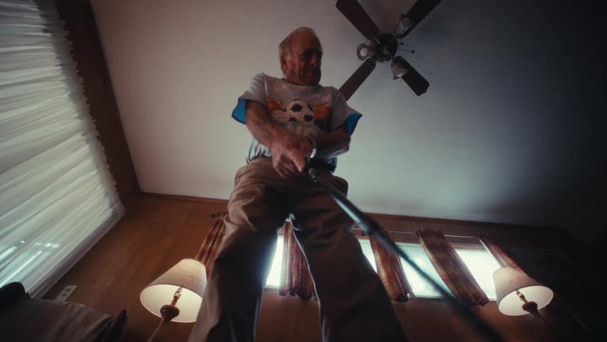 An elderly man in a soccer graphic tshirt plays minigolf filmed from a low angle looking up as he moves the stick back and forth beneath a wooden ceiling fan and in front of woodpaneled walls with two small table lamps