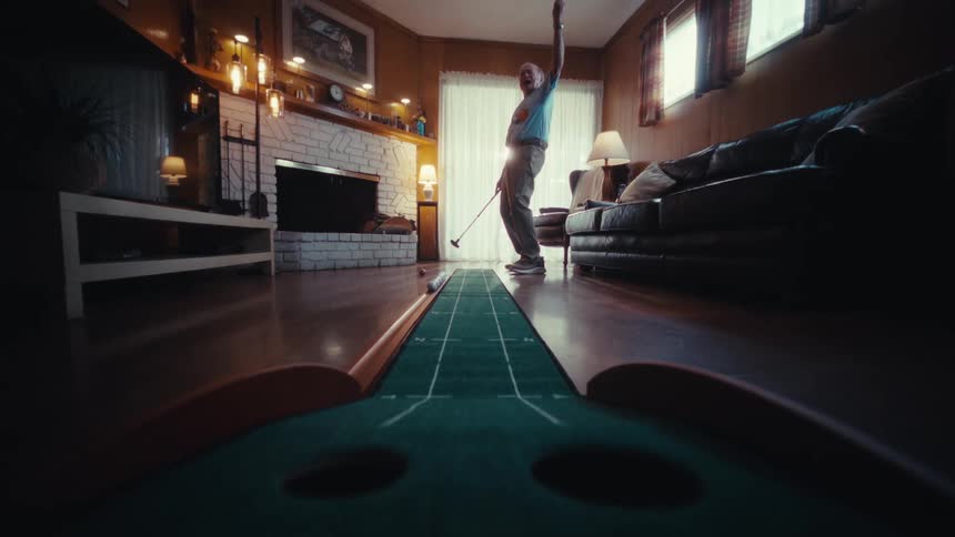 Man standing in his living room holding a golf club as he putts a ball into the hole and cheers