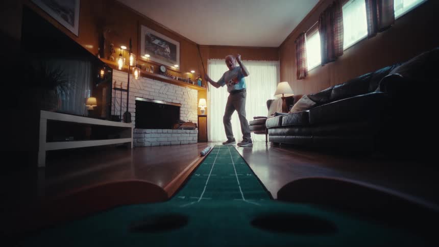 Man standing in his living room holding a golf club as he putts a ball into the hole and cheers