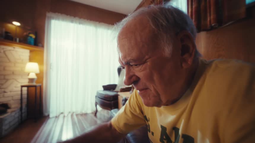 An elderly man wearing a yellow us army veteran tshirt sits on a leather sofa and opens a laptop on the coffee table