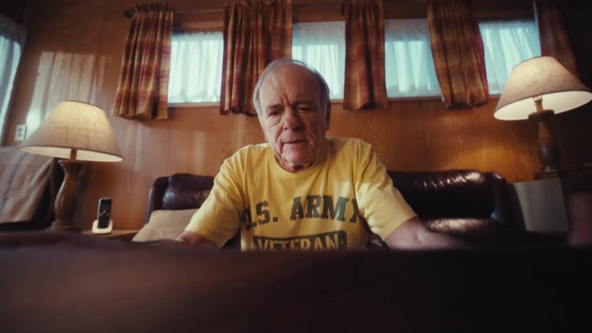 An elderly man wearing a yellow us army veteran tshirt sits on a leather sofa in a woodpaneled living room while looking down at a laptop screen on a table