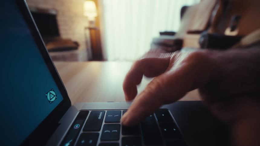 A person uses a laptop on a wooden table in a dimly lit living room focusing on their hand as they type on the keyboard and move their fingers across the trackpad while a screen glows in the foreground