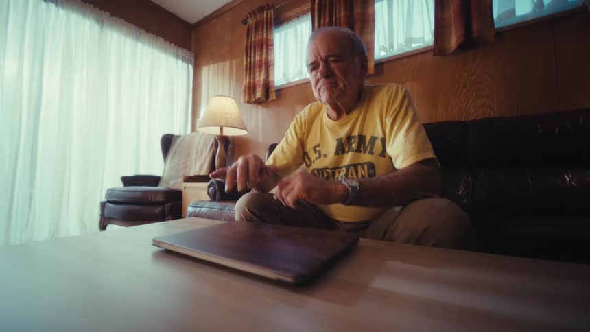 An elderly man sitting in his living room at midday wearing a yellow veteran tshirt closing his laptop lid in disappointment