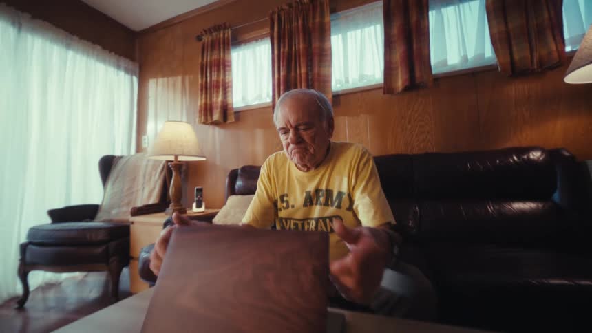 An elderly army veteran sits on a couch and opens a laptop in his living room