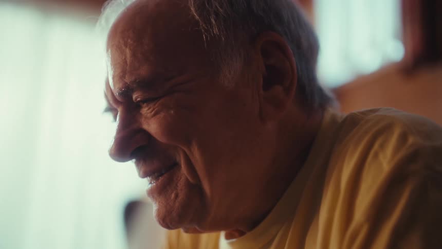 An elderly man in a yellow shirt sits in a sunlit room laughing heartily and then settling into a gentle smile while looking down