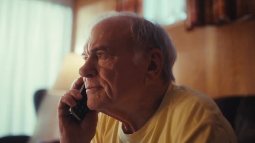 Man sitting on his living room couch typing on a laptop computer when he answers his phone and gets excited